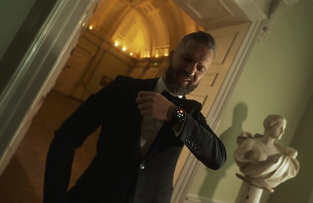 Mężczyzna w garniturze patrzy na smartwatch na swoim nadgarstku w eleganckim wnętrzu z rzeźbą w tle. Smartwatch ma okrągły wyświetlacz z kolorowym interfejsem.
