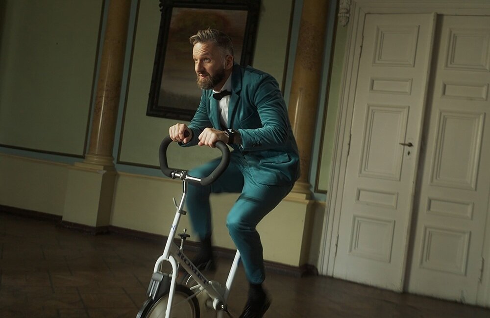 Mężczyzna w eleganckim garniturze jedzie na rowerze stacjonarnym w pomieszczeniu z klasycznym wystrojem. Na jego nadgarstku widoczny jest smartwatch.