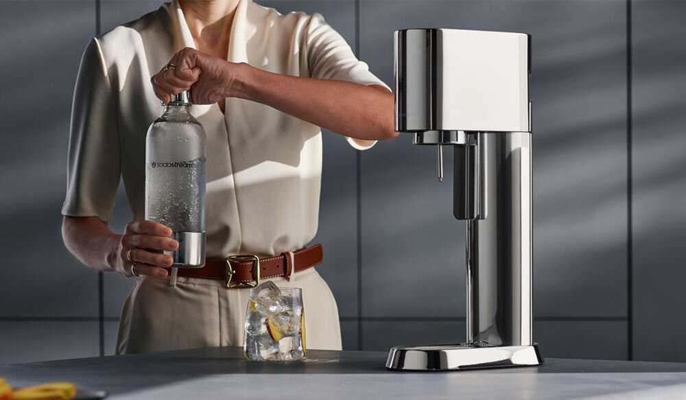 Osoba trzyma butelkę z wodą gazowaną obok stalowego saturatora. Na butelce widoczny jest napis \'sodastream\'.
