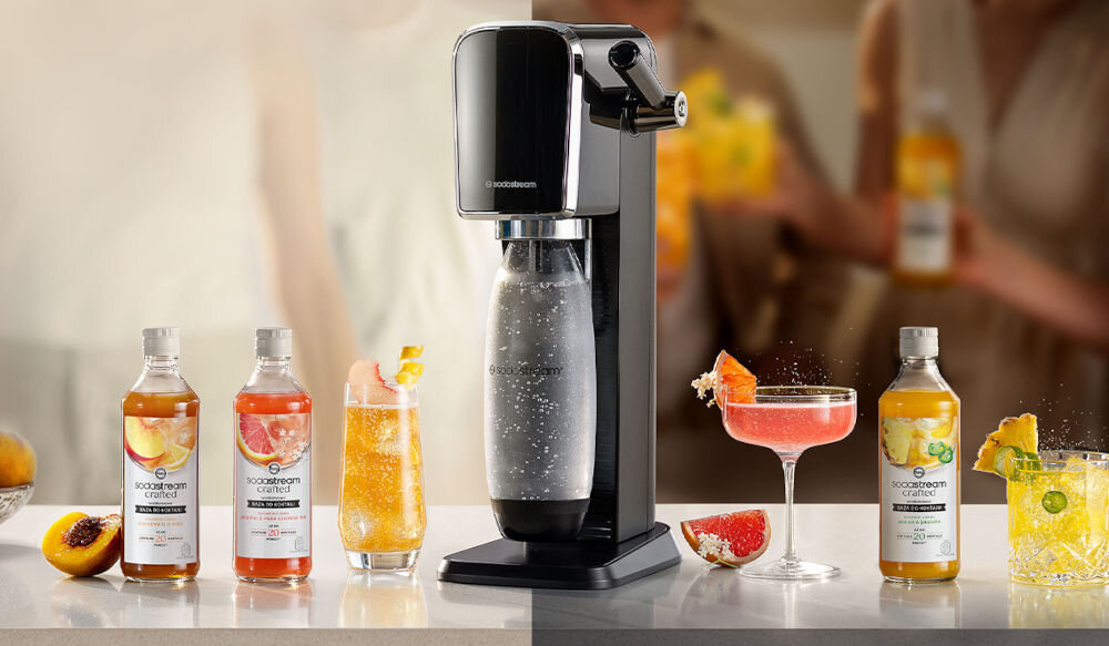 Na blacie stoi saturator z logo \'sodastream\', obok którego znajdują się cztery butelki z syropami oraz trzy szklanki z napojami. W tle widoczne są rozmyte sylwetki osób.