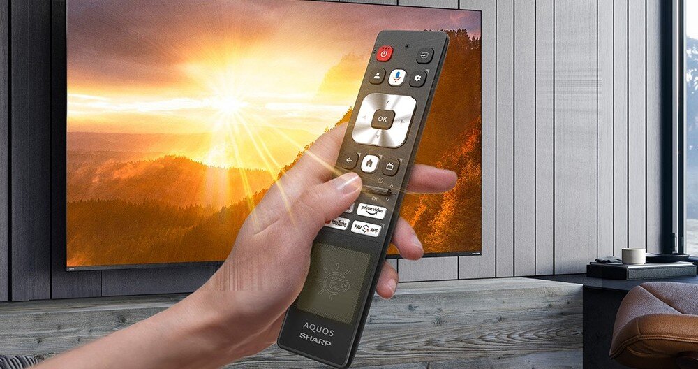 Telewizor SHARP 55JT8765E z pilotem solarnym, zasilanie energią światła i możliwość ładowania przez port USB-C, telewizor zawieszony na ścianie w pomieszczeniu, pilot trzymany w ręku