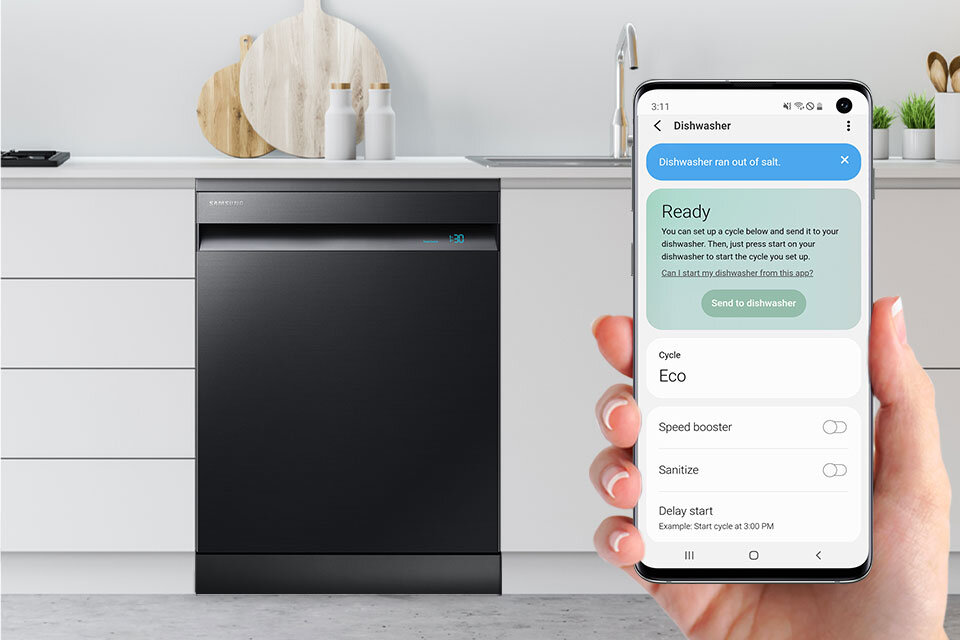 Aplikacja SmartThings Czarna zmywarka SAMSUNG w zabudowie kuchennej z widocznym logo producenta. Obok, osoba trzyma smartfon z aplikacją pokazującą status zmywarki: \'Dishwasher ran out of salt\', \'Ready\', \'Cycle Eco\'.