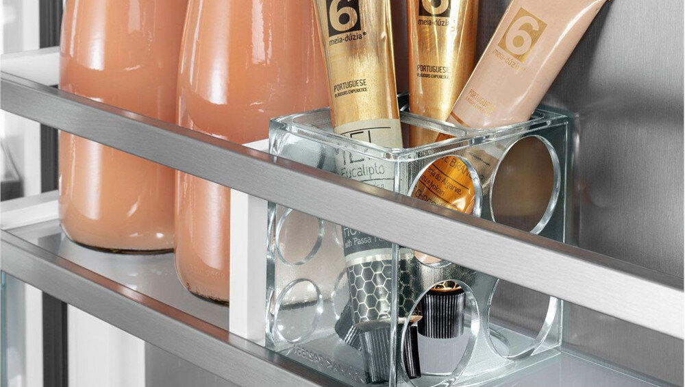 Na półce znajdują się butelki z różowym płynem oraz przezroczysty pojemnik z tubkami kosmetyków. Widoczne są napisy na tubkach, w tym \'6\' i \'PORTUGUESE\'.