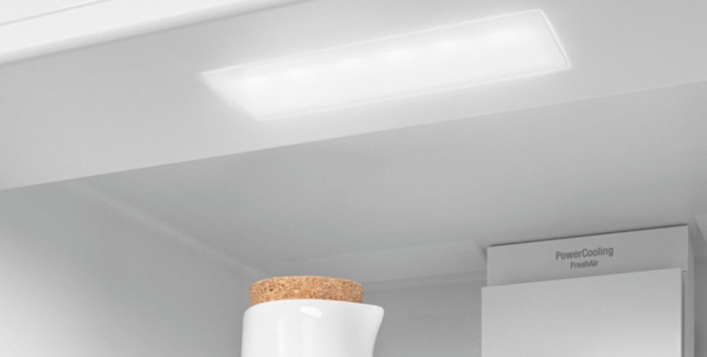 Wnętrze lodówki z widocznym oświetleniem LED i fragmentem białego pojemnika z korkową pokrywką. W tle napis \'PowerCooling FreshAir\'.