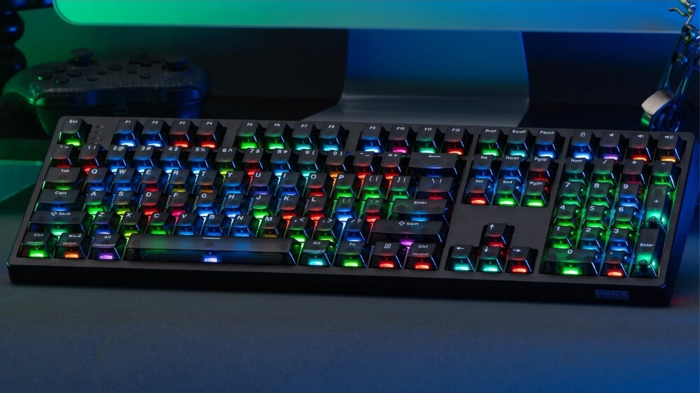 Klawiatura mechaniczna z podświetleniem RGB, z widocznymi różnokolorowymi klawiszami na ciemnym tle. W tle znajduje się monitor i kontroler do gier.