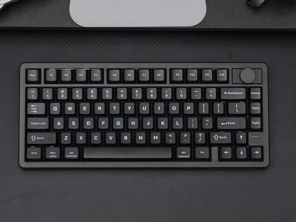 Czarna klawiatura mechaniczna z układem QWERTY, posiadająca klawisze funkcyjne od F1 do F12 oraz pokrętło w prawym górnym rogu. Widoczne są klawisze takie jak Esc, Tab, Caps Lock, Shift, Ctrl, Alt, Enter, Backspace, Delete, PgUp, PgDn oraz strzałki kierunkowe.