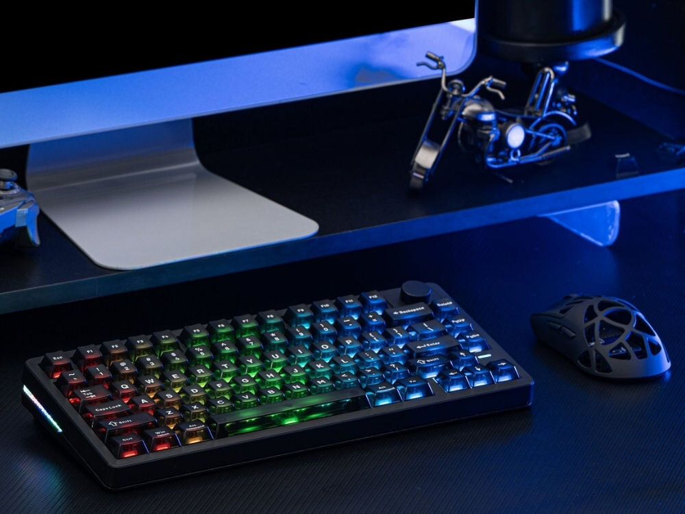 Na biurku znajduje się czarna klawiatura z podświetleniem RGB, obok niej mysz komputerowa. W tle widoczny jest monitor oraz metalowy model motocykla.