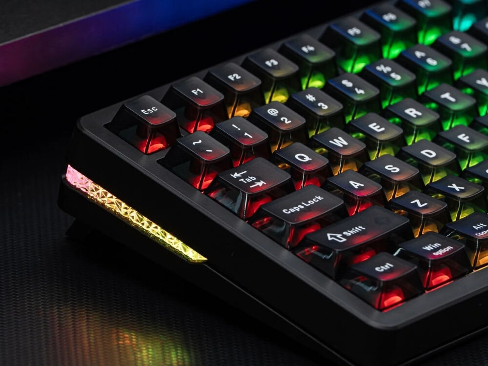 Czarna klawiatura mechaniczna z podświetleniem RGB, widoczne klawisze to m.in. Esc, F1-F12, QWERTY, CapsLock, Shift, Ctrl. Boczna krawędź klawiatury ma dodatkowe podświetlenie w kolorach tęczy.