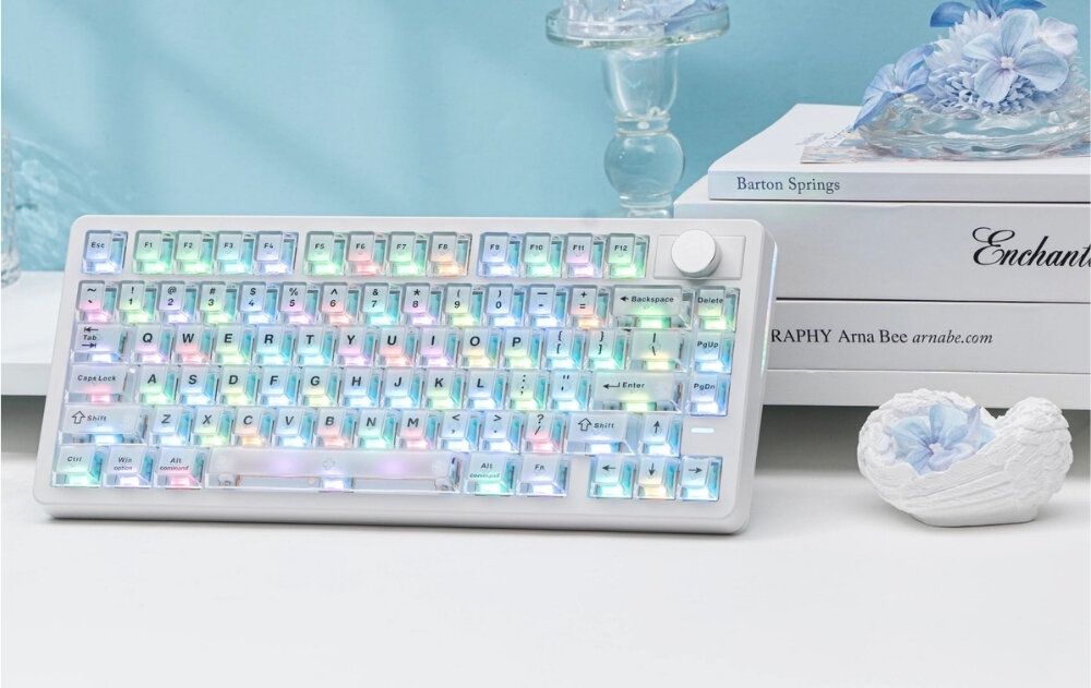 Biała klawiatura z podświetleniem RGB, z pokrętłem w prawym górnym rogu. W tle widoczne są książki i dekoracje w jasnych kolorach.