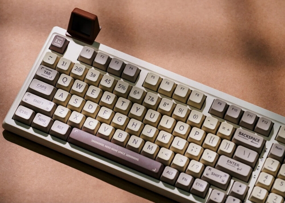 Klawiatura mechaniczna w stylu retro z białymi i beżowymi klawiszami, na brązowym tle. Widoczny jest napis na klawiszu spacji: \'NEED MORE SPACE\'.
