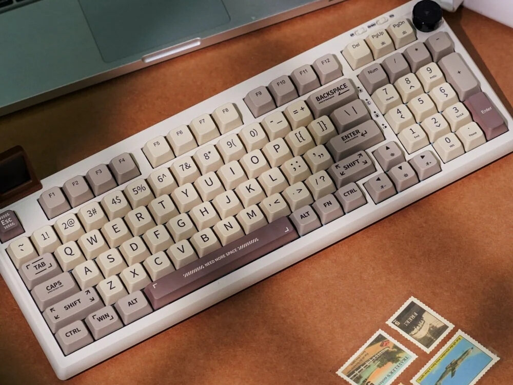 Na obrazie widoczna jest klawiatura w stylu retro z kremowymi i brązowymi klawiszami, na drewnianym biurku. Obok klawiatury znajdują się znaczki pocztowe i fragment laptopa.