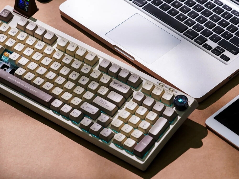 Na obrazie widoczna jest klawiatura o retro wyglądzie z jasnymi klawiszami i ciemniejszymi akcentami, umieszczona obok laptopa. Na klawiaturze widoczny jest napis \'BACKSPACE\'.