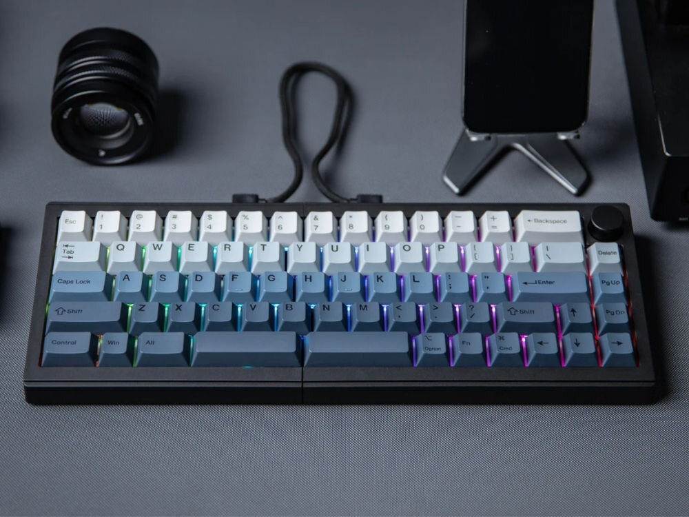 Na obrazie widoczna jest klawiatura mechaniczna z podświetleniem RGB, z klawiszami w odcieniach szarości i bieli. W tle znajdują się obiekty takie jak obiektyw aparatu i stojak na telefon.