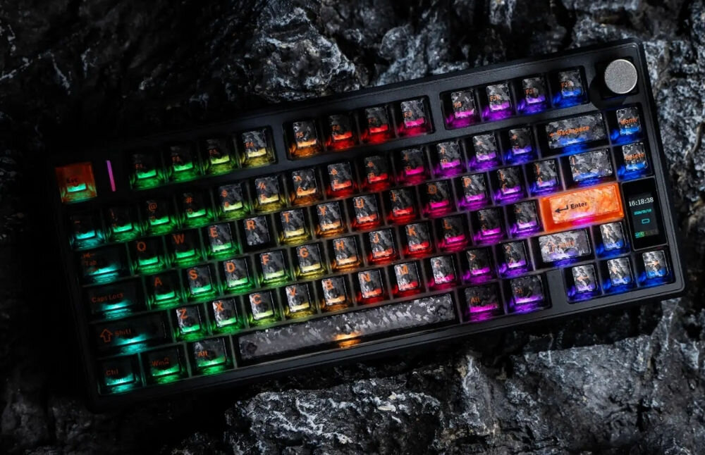 Czarna klawiatura mechaniczna z podświetleniem RGB, leżąca na ciemnym, kamienistym tle. Klawisze są podświetlone w różnych kolorach, a na jednym z nich widoczny jest napis \'Enter\'.