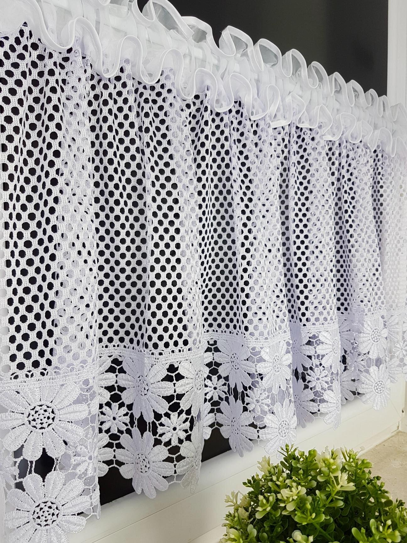Białe firany z siateczki z gipiurowym wzorem stokrotek, zawieszone na karniszu. Na parapecie widoczna jest zielona roślina doniczkowa.