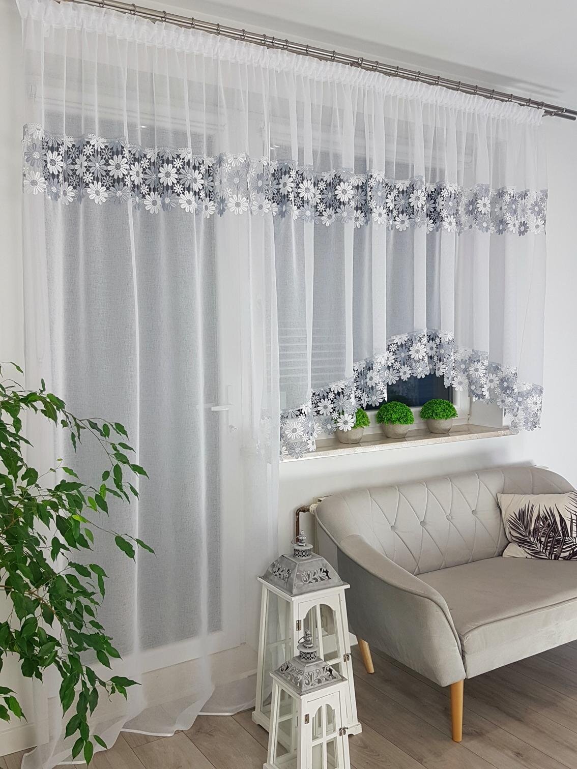 Na zdjęciu widoczny jest komplet białych firan balkonowych z szarą gipiurą, zawieszony na karniszu w jasnym wnętrzu. Pod oknem znajduje się sofa, a na parapecie ustawione są zielone rośliny.
