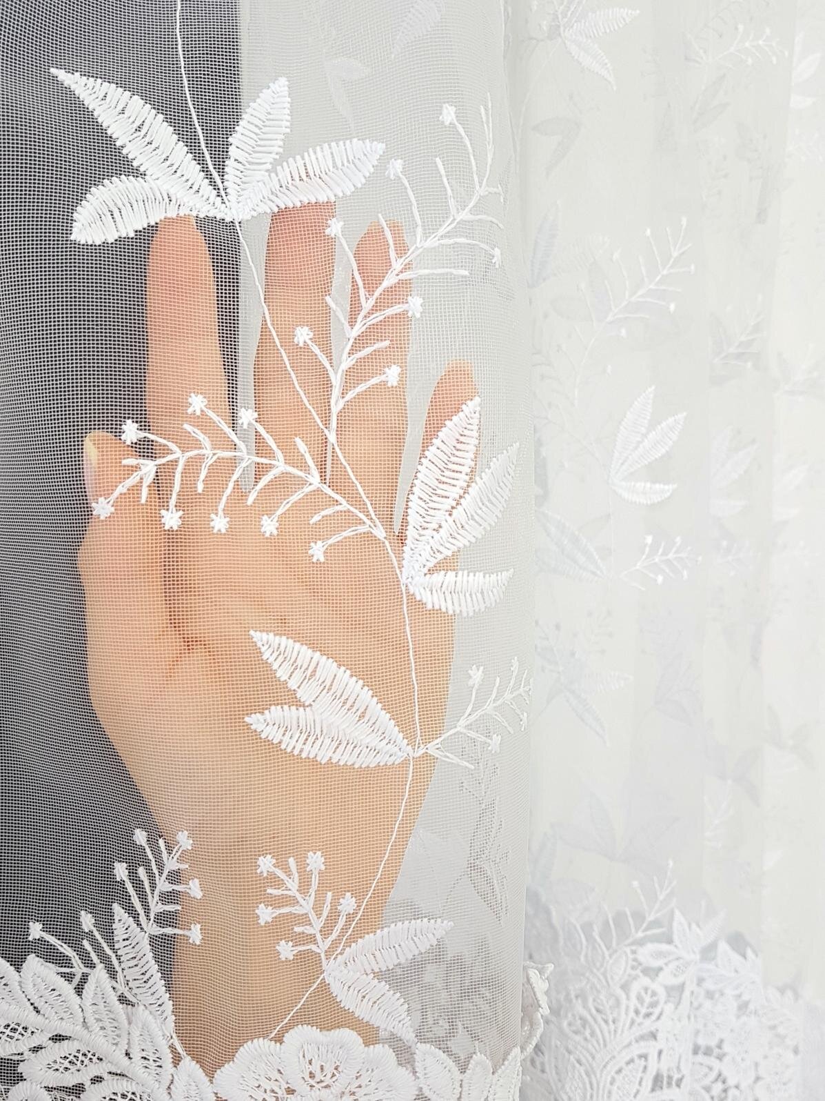 Ręka dotyka białej firany z markizety z haftowanym wzorem listków i kwiatów. Firana ma delikatny, ażurowy wzór z gipiurą na dole.