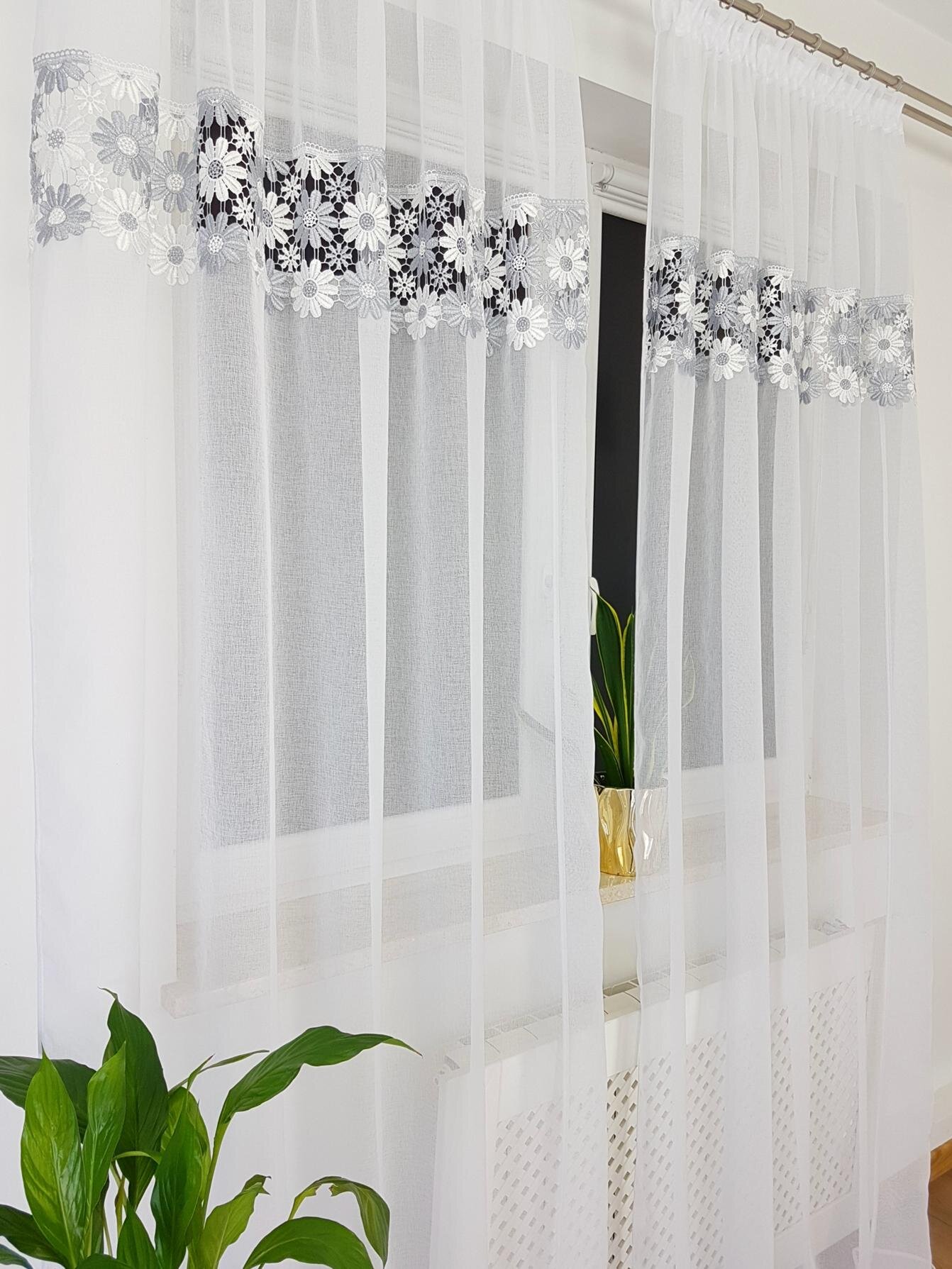 Na zdjęciu widać białą firanę balkonową z podwójną gipiurą w kwiatowy wzór, zawieszoną na karniszu. W tle znajduje się okno oraz roślina doniczkowa na parapecie.