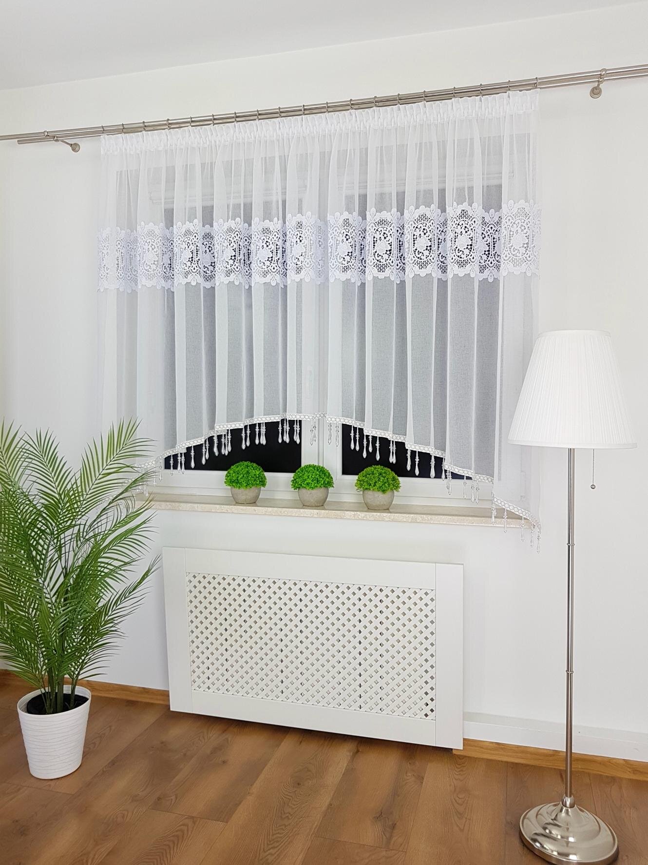 Na zdjęciu widać elegancką firanę z gipiurą i koralikami zawieszoną na karniszu nad oknem. W pomieszczeniu znajduje się również lampa stojąca, roślina doniczkowa i trzy małe rośliny na parapecie.