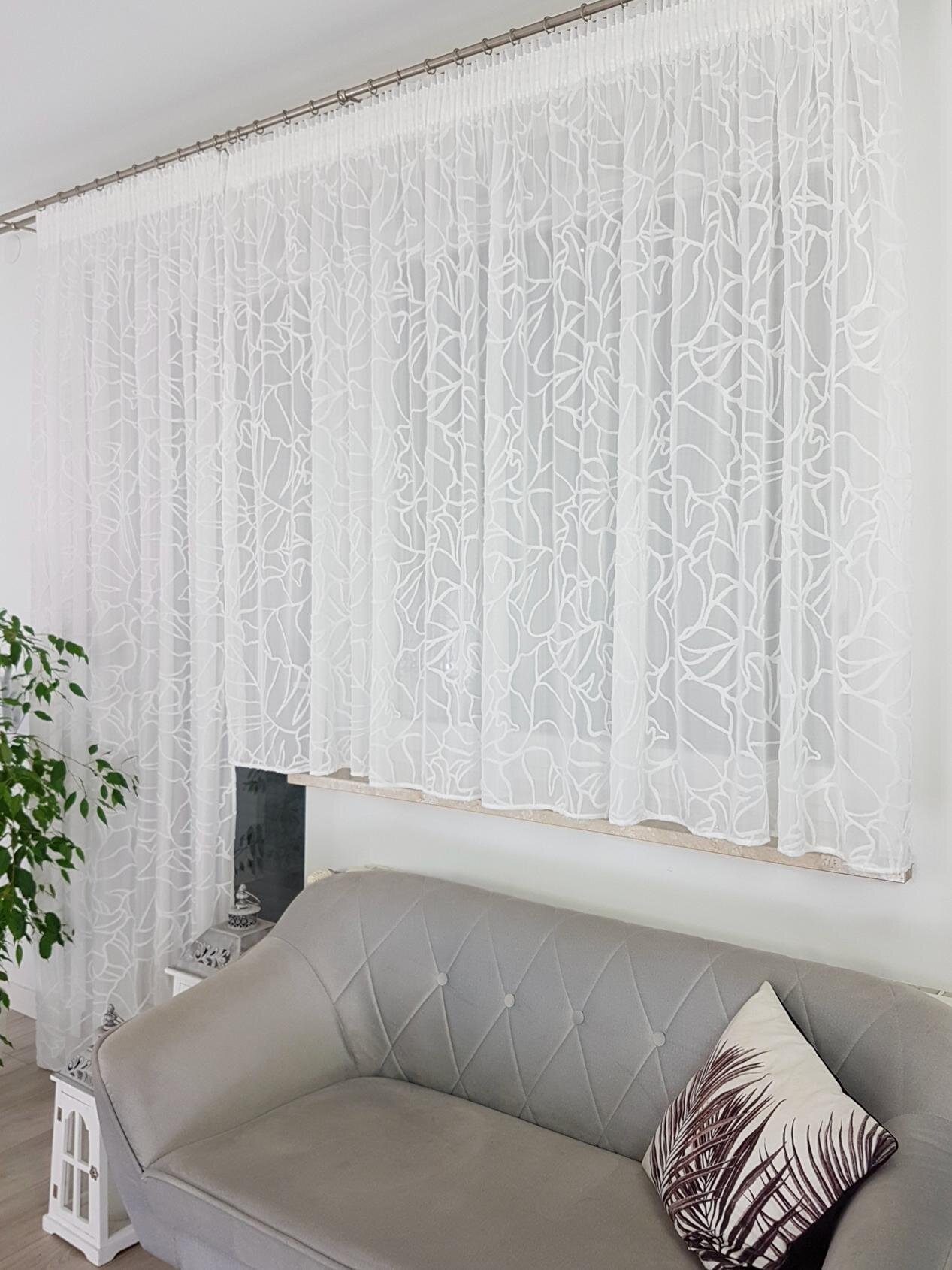 Na zdjęciu widoczny jest komplet białych firan z reliefowym wzorem 3D zawieszonych na karniszu. Przed firanami znajduje się szara sofa z poduszką ozdobną, a obok stoi roślina w doniczce.