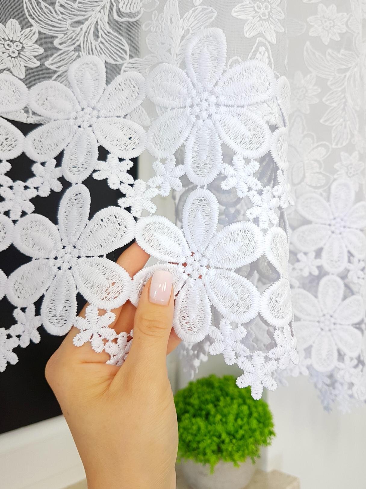 Biała firana z haftem 3D przedstawiającym duże kwiaty, trzymana przez dłoń z jasnym lakierem na paznokciach. W tle widoczna jest zielona roślina w doniczce.