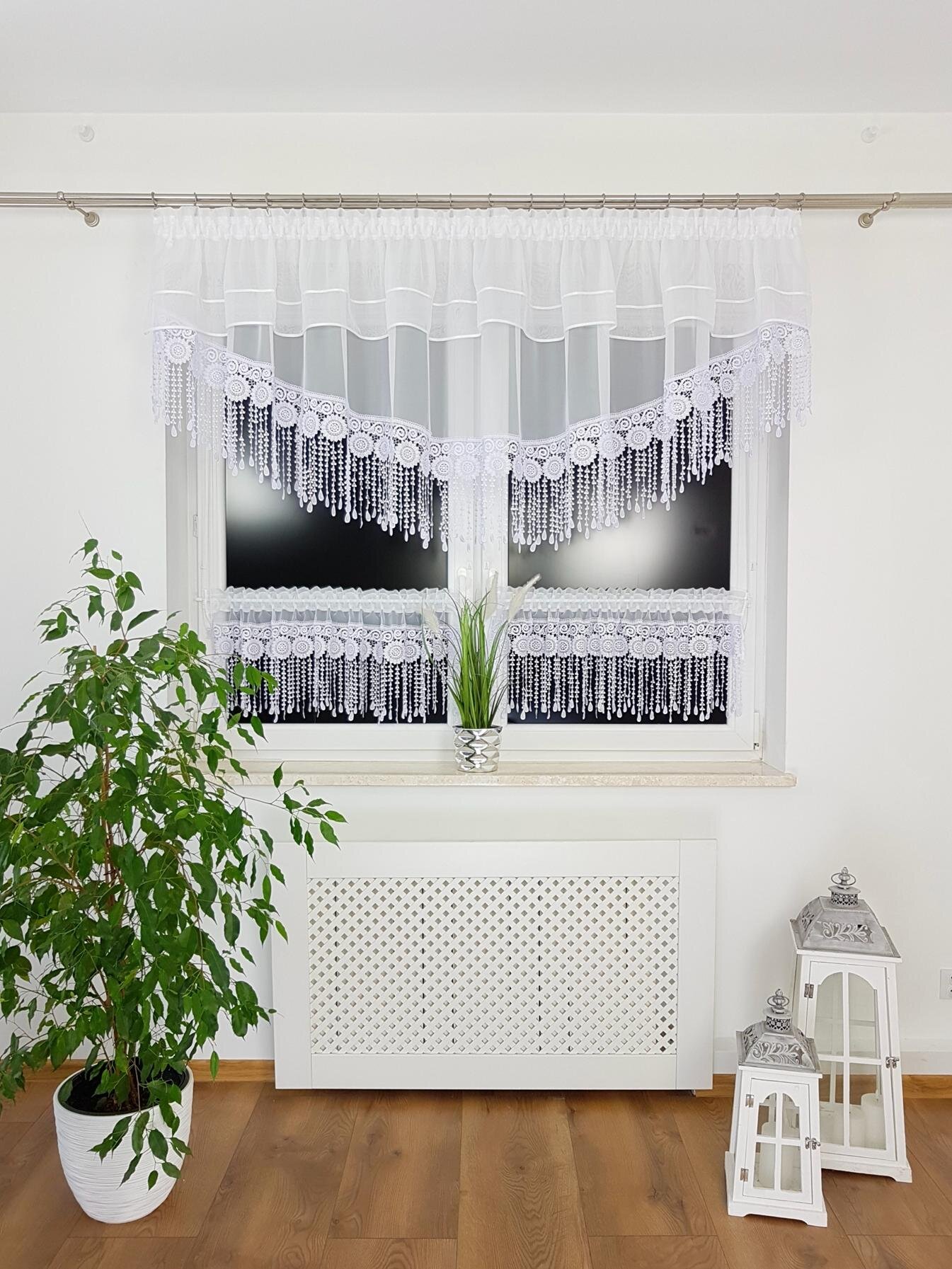 Na zdjęciu znajduje się okno ozdobione białą firaną z zazdrostkami, wykonane z woalu, lambrekinu i gipiury. Przed oknem stoi roślina doniczkowa, a obok znajdują się dwie dekoracyjne latarnie.