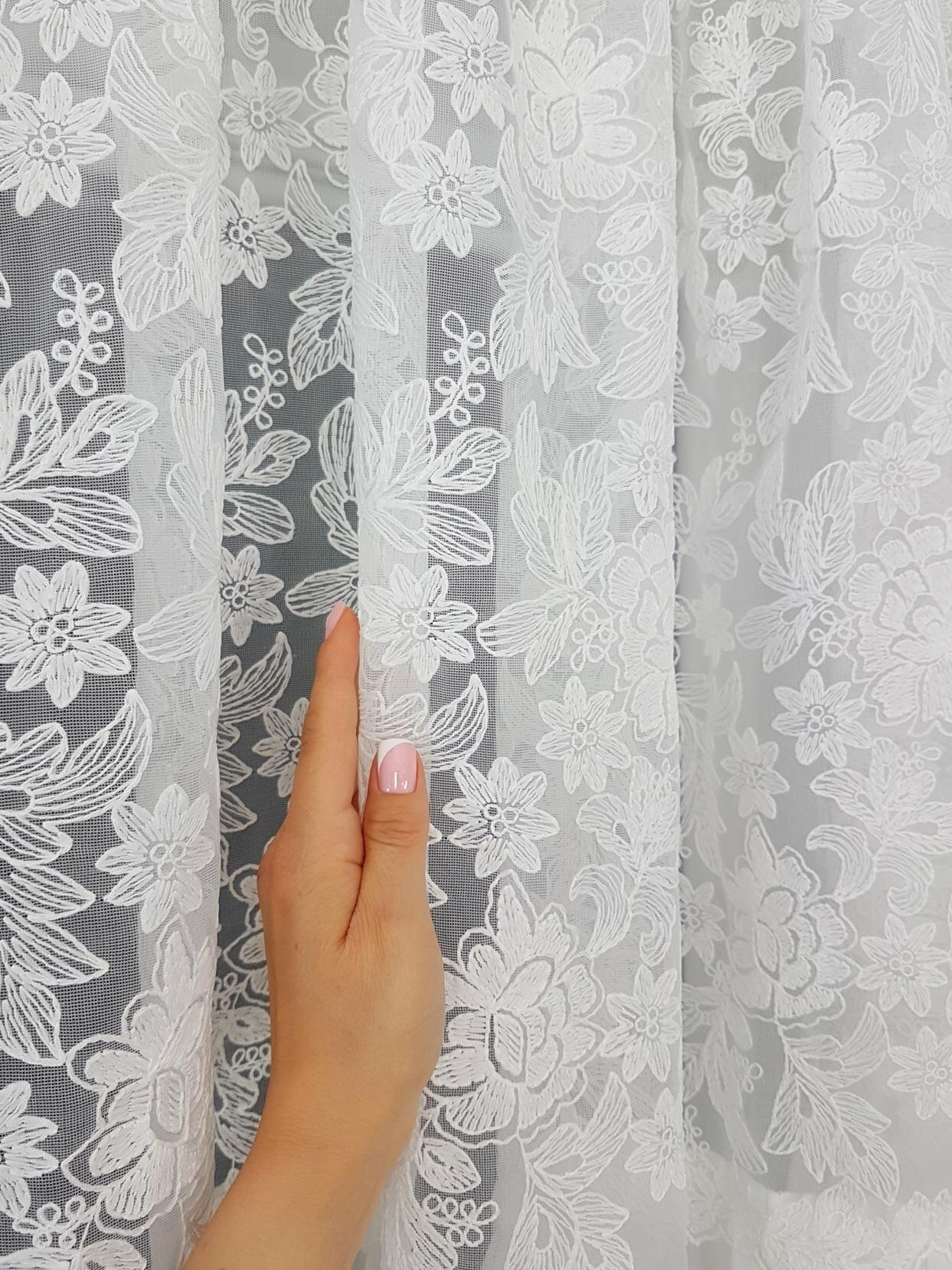 Ręka dotyka białej firany z haftowanym wzorem kwiatowym. Tkanina jest półprzezroczysta, z wyraźnym, delikatnym wzorem roślinnym.