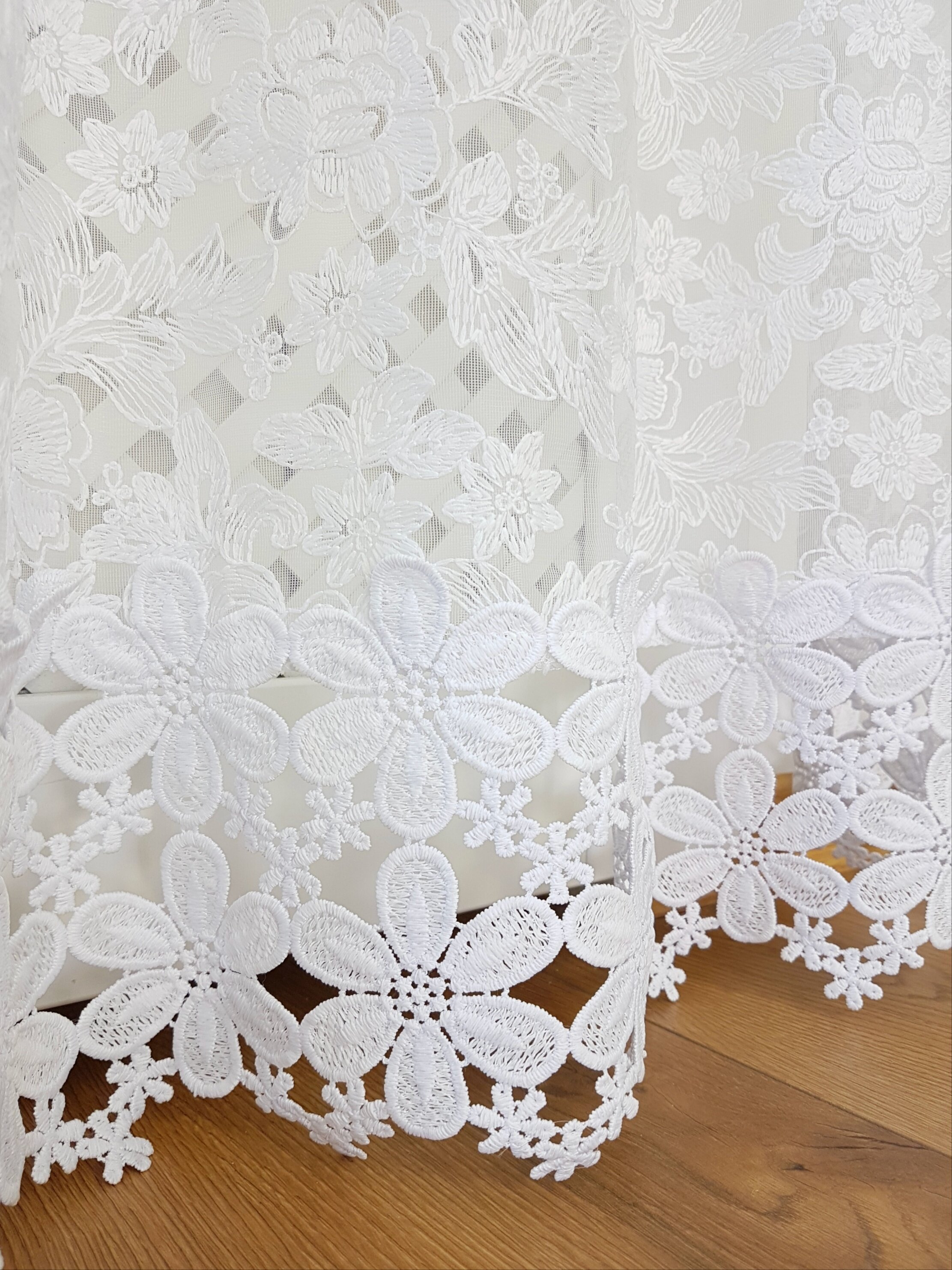 Biała firana z haftem 3D przedstawiającym kwiaty i gipiurą na dole. Tło jest drewniane, co podkreśla delikatność materiału.