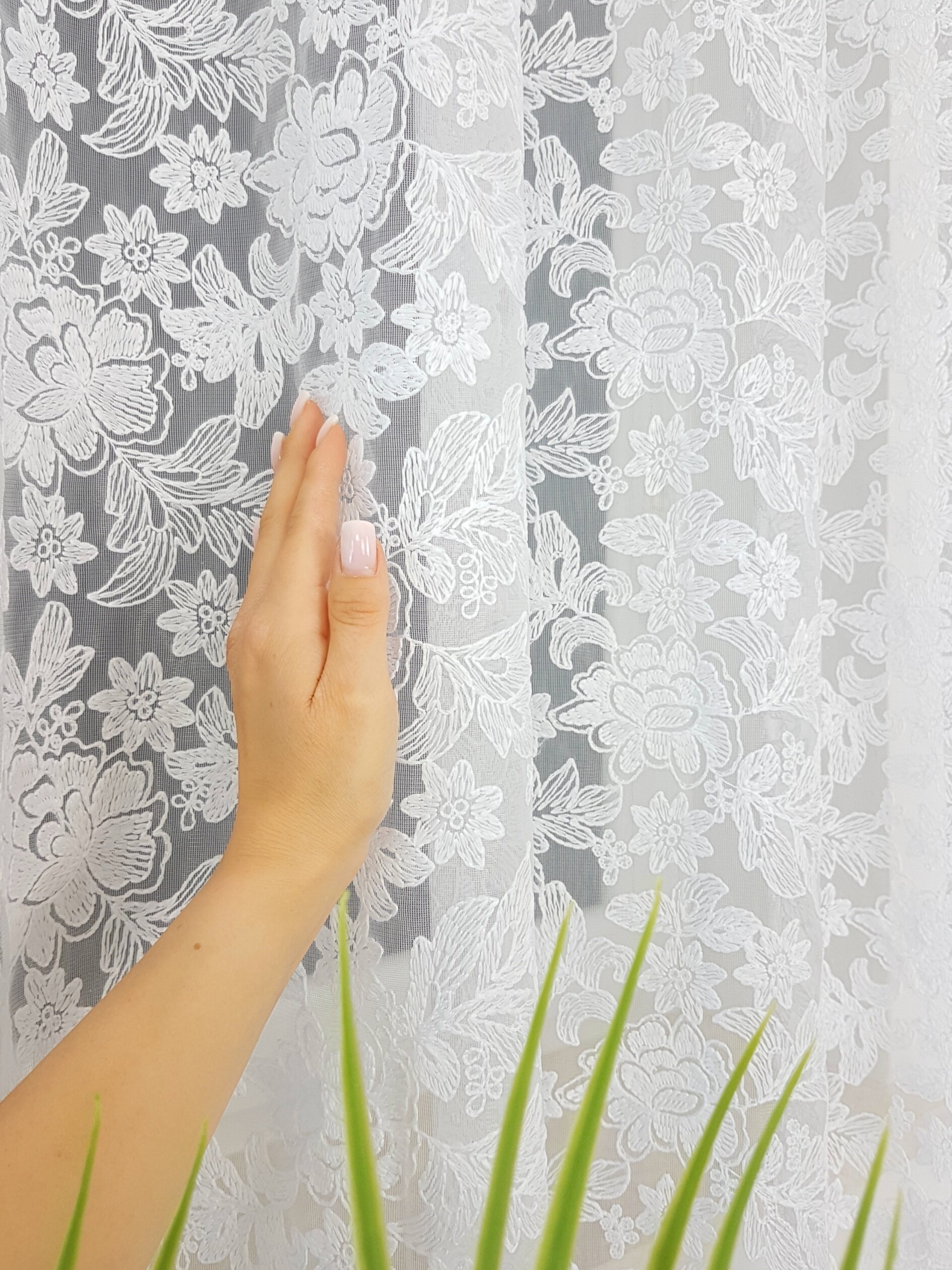 Biała firana z haftem 3D i gipiurą, na której widoczny jest wzór kwiatowy. Obok firany znajduje się dłoń oraz fragment zielonej rośliny.