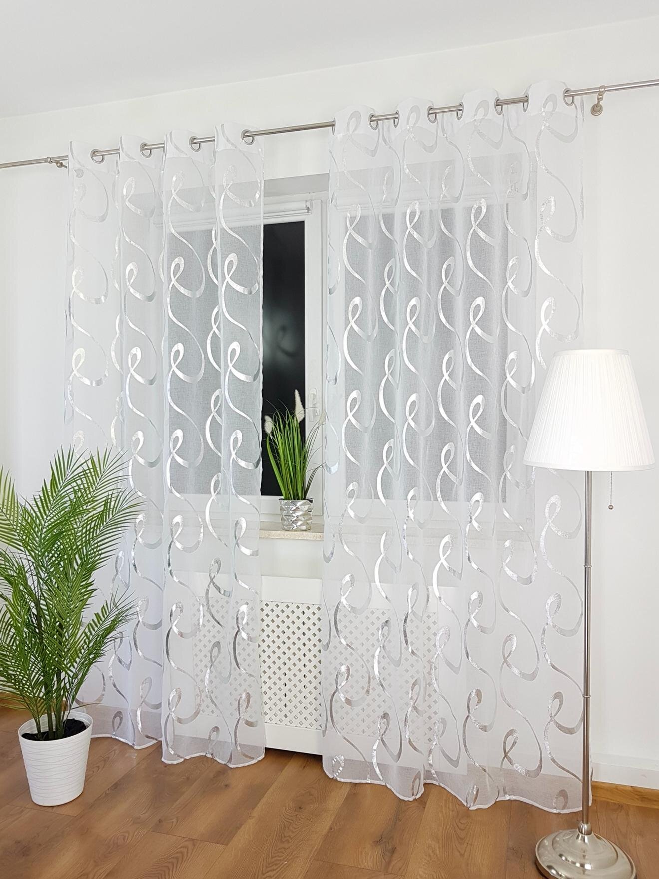 Na zdjęciu widać okno z zawieszoną firaną z etaminy, uszytą na przelotkach, z wzorem w srebrne spirale. Obok okna stoi lampa podłogowa i doniczka z rośliną.