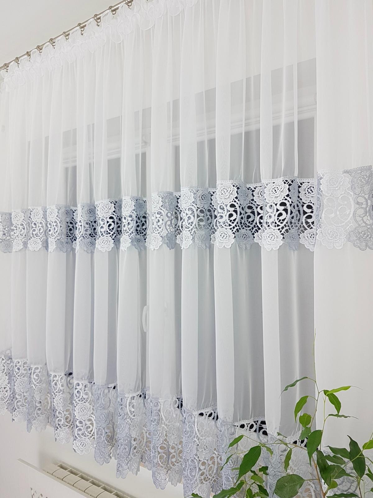 Firana z woalu o wymiarach 400x180 cm, ozdobiona szerokimi gipiurami w dwóch poziomych pasach. W tle widoczne jest okno oraz fragment rośliny doniczkowej.