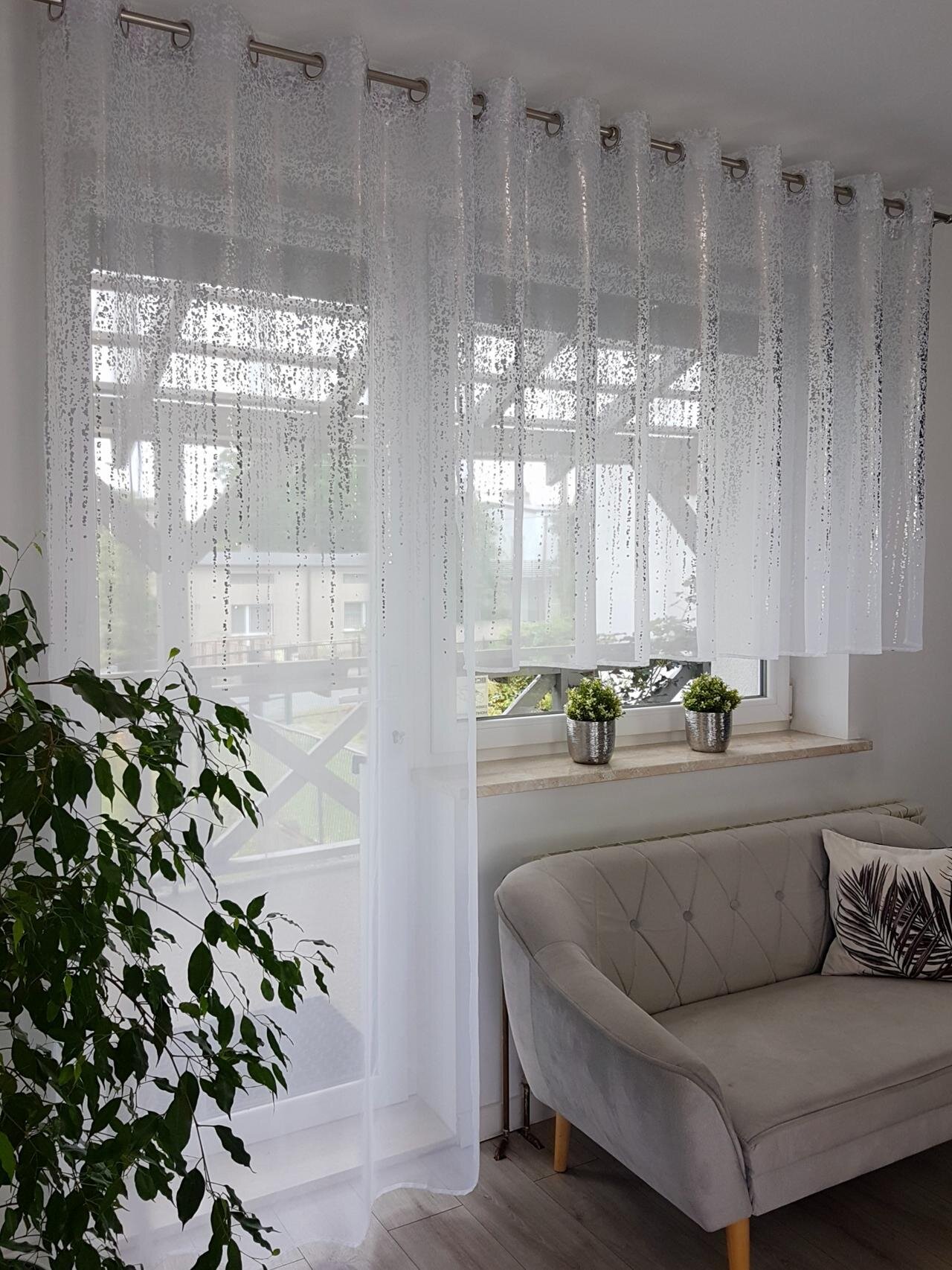 Na obrazie widoczny jest komplet białych firan z woalu z wzorem przypominającym srebrny deszczyk, zawieszony na przelotkach. W pomieszczeniu znajduje się szara sofa, roślina doniczkowa oraz trzy małe rośliny na parapecie.