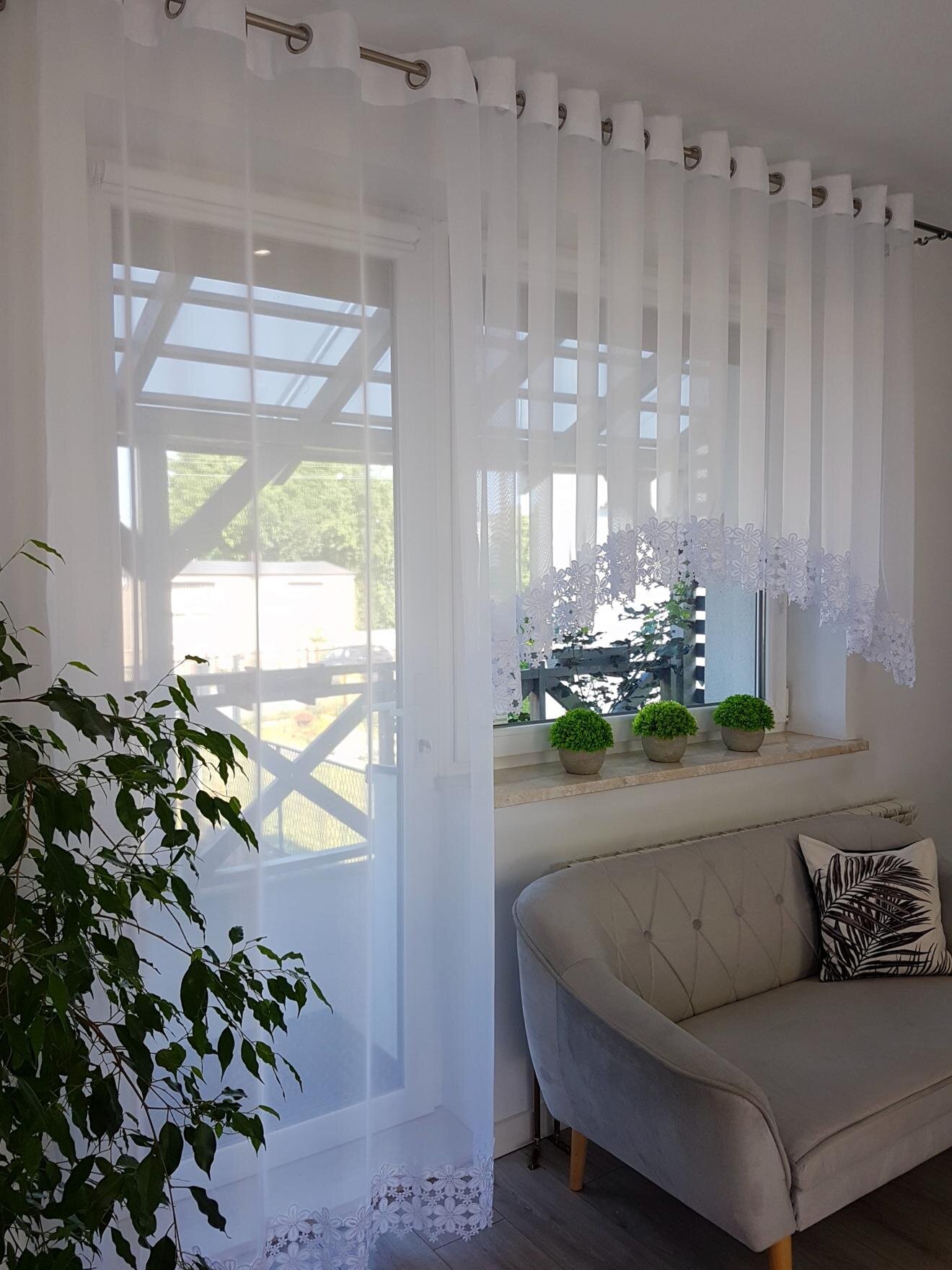 Białe zasłony z woalu na przelotkach, zdobione gipiurą, wiszą w oknie. Na parapecie stoją trzy małe rośliny w doniczkach, a obok znajduje się jasna sofa z poduszką.