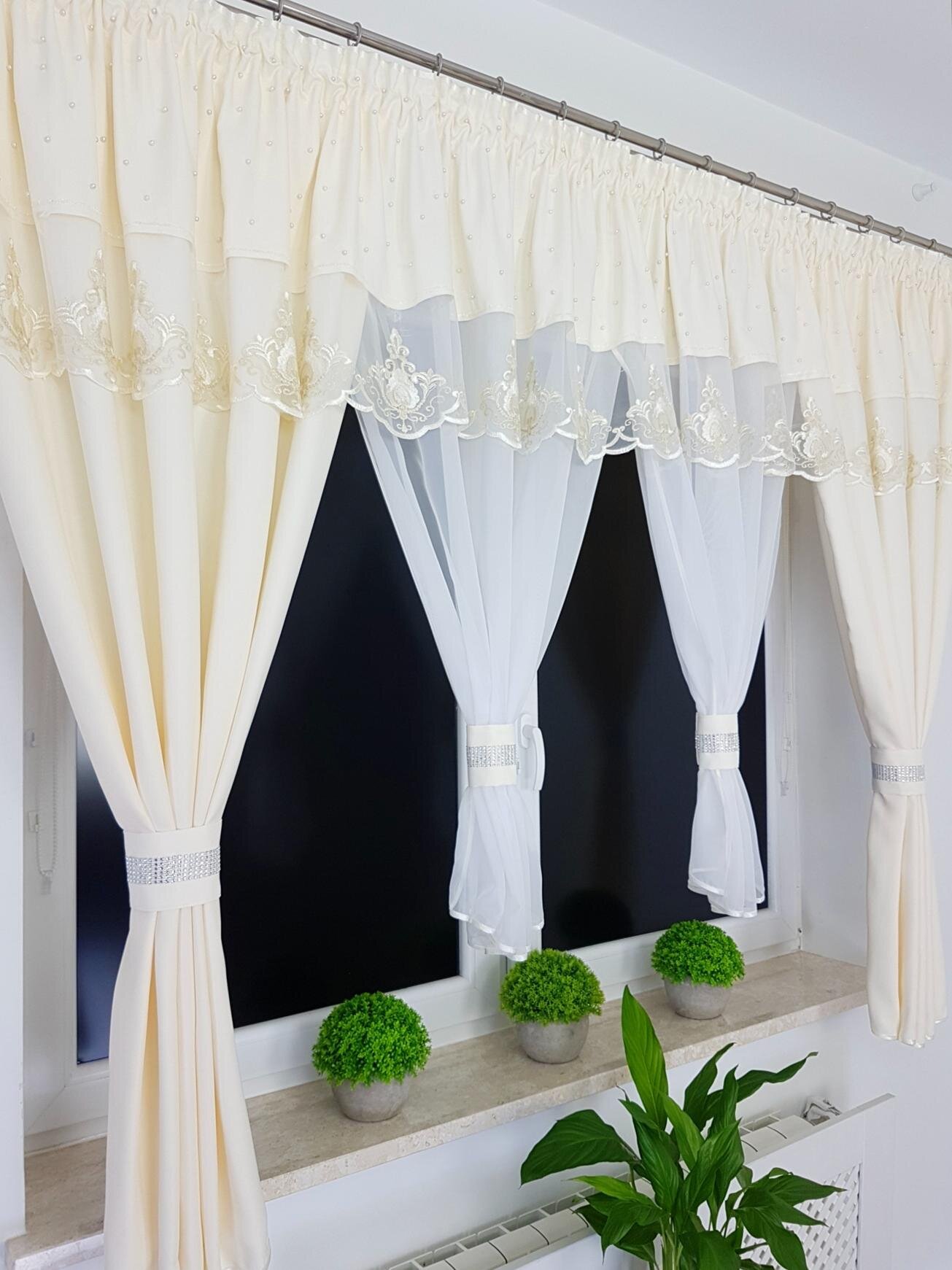 Na zdjęciu widać kremowe firany z lambrekinem, ozdobione delikatnym haftem, zawieszone na karniszu. Na parapecie znajdują się trzy doniczki z zielonymi roślinami.