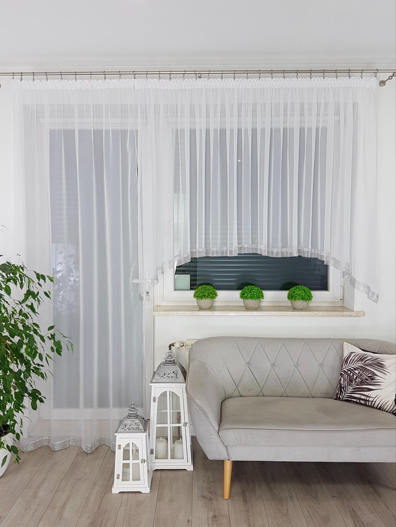 Wnętrze pokoju z białymi firanami na oknie i drzwiach balkonowych, obok których stoi szara sofa z poduszką. Na parapecie znajdują się trzy małe rośliny w doniczkach, a obok sofy stoją dwie białe latarnie dekoracyjne.