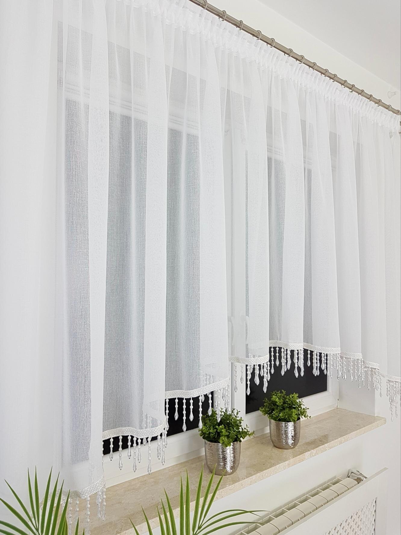 Na zdjęciu widać białe firany z etaminy zawieszone na karniszu, ozdobione koralikami na dole. Pod oknem znajdują się trzy srebrne doniczki z zielonymi roślinami.