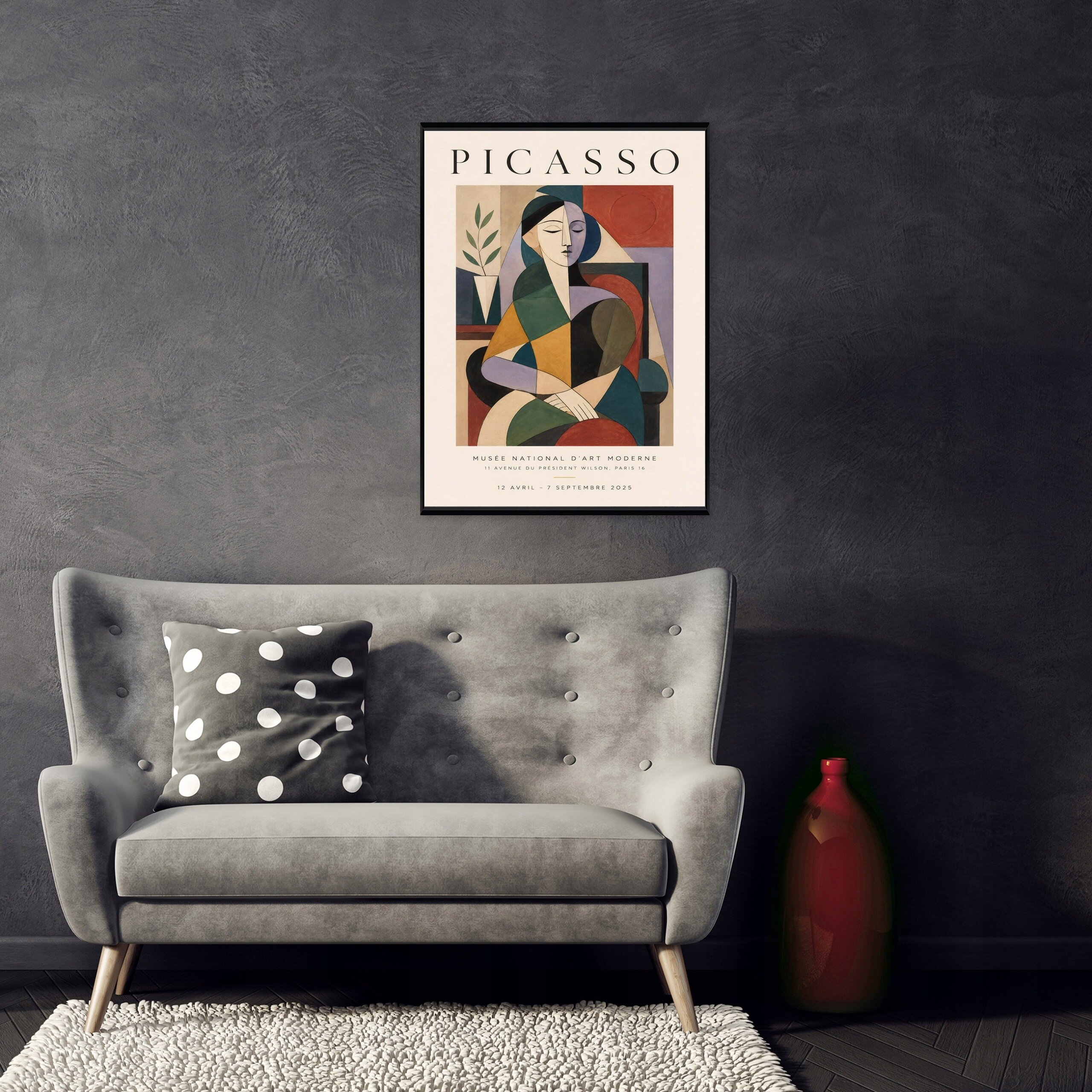 Na ścianie wisi obraz z napisem \'PICASSO\', przedstawiający abstrakcyjną postać w kolorach ziemi. Przed obrazem stoi szara sofa z poduszką w białe kropki, a obok niej znajduje się czerwona waza.
