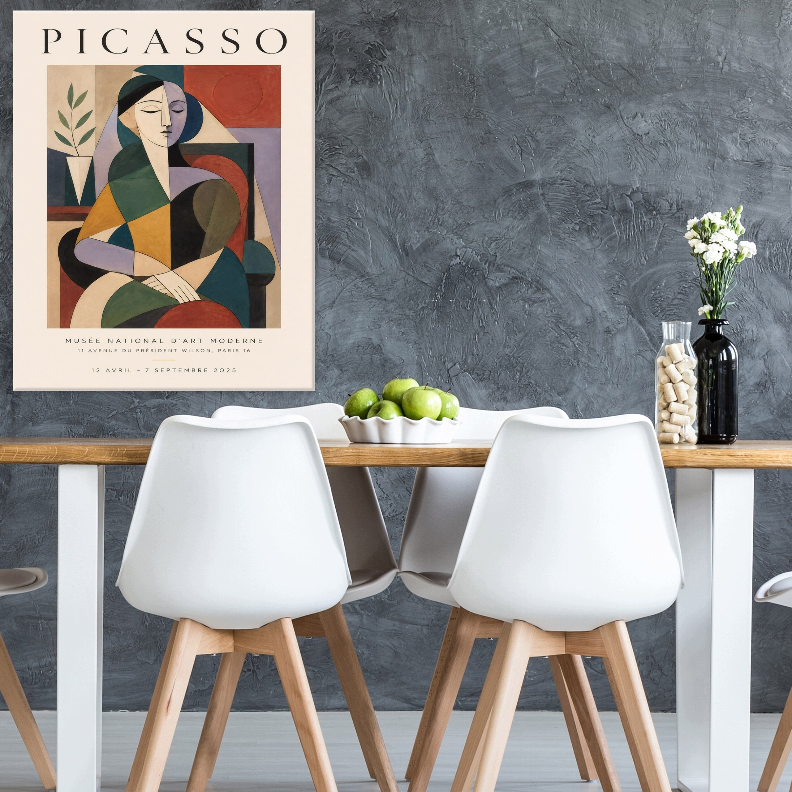 Na ścianie wisi plakat z napisem \'PICASSO\' i abstrakcyjnym obrazem przedstawiającym postać siedzącą. Przed plakatem stoi stół z białymi krzesłami, na którym znajduje się misa z zielonymi jabłkami oraz wazon z kwiatami.