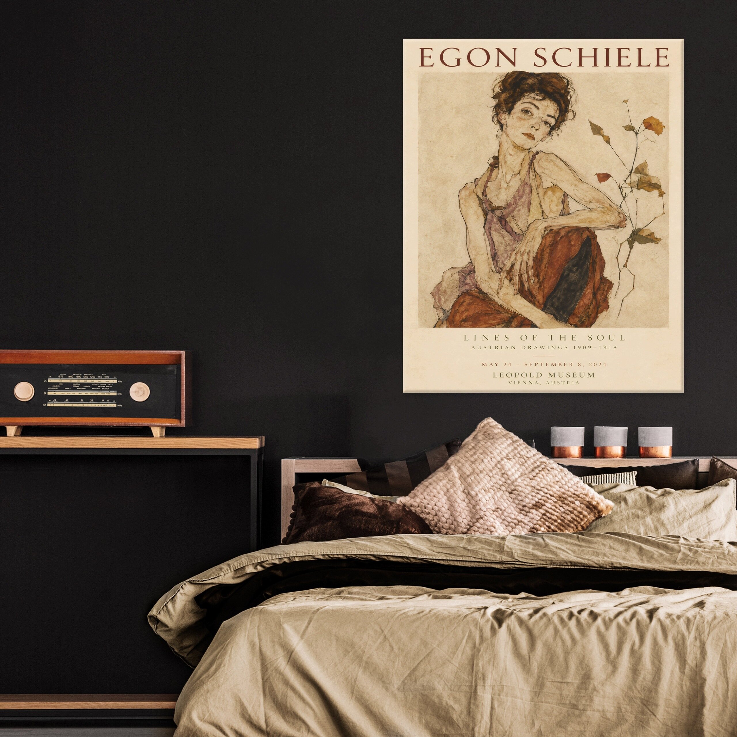 Na ścianie wisi obraz przedstawiający kobietę, z tekstem \'EGON SCHIELE LINES OF THE SOUL\'. Obok znajduje się drewniana półka z radiem i kilka poduszek na łóżku.