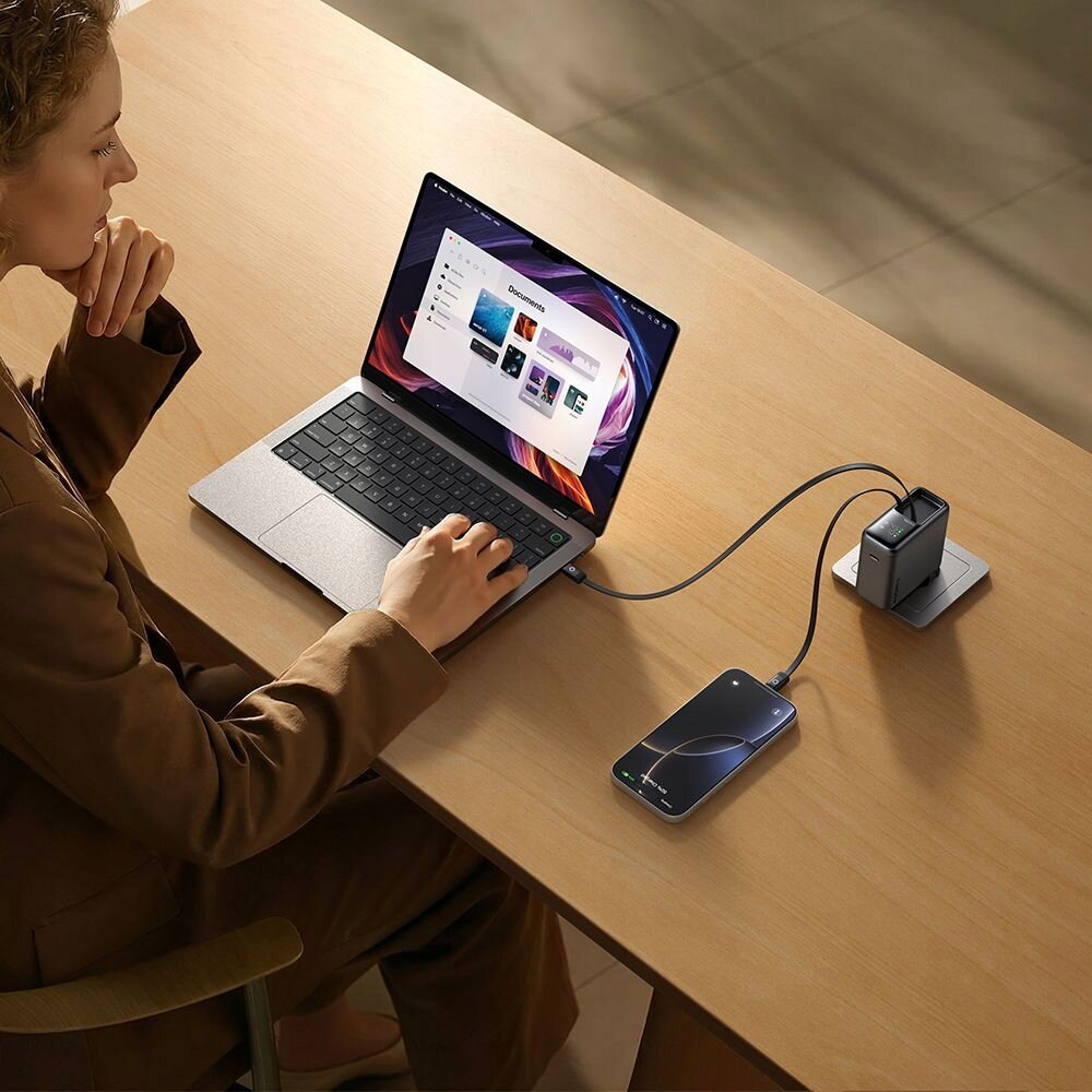 Osoba siedzi przy drewnianym stole, używając laptopa, który jest podłączony do ładowarki sieciowej. Obok laptopa znajduje się smartfon również podłączony do ładowarki za pomocą kabla.