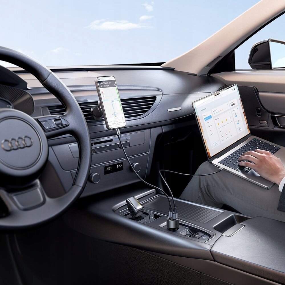 Wnętrze samochodu z widocznym logo Audi na kierownicy. Telefon i laptop są podłączone do ładowarki samochodowej umieszczonej w konsoli środkowej.