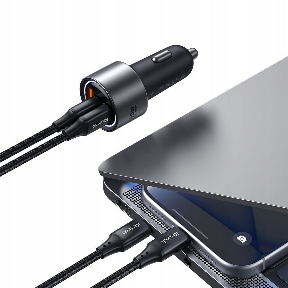 Na obrazie widoczna jest ładowarka samochodowa z trzema podłączonymi kablami USB, obok laptopa i smartfona. Na kablach widoczny jest napis \'Mcdodo\'.