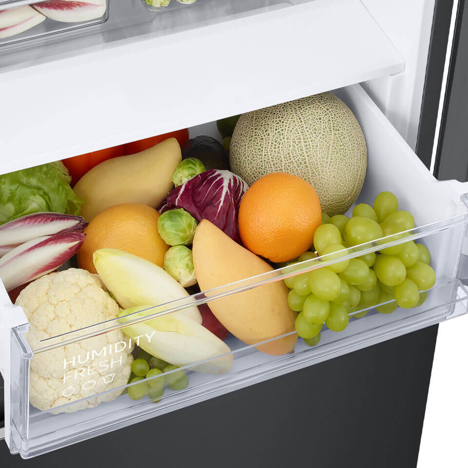 Specjalna szuflada na owoce i warzywa Szuflada lodówki wypełniona różnorodnymi owocami i warzywami, takimi jak winogrona, melon, pomarańcza, sałata i kalafior. Na szufladzie widoczny jest napis \'HUMIDITY FRESH\'.