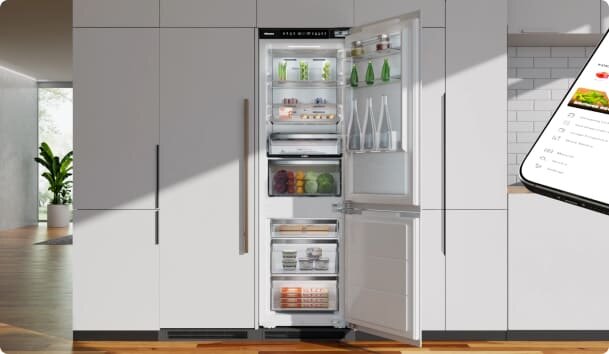Otwarta lodówka z widocznymi półkami, na których znajdują się różne produkty spożywcze, takie jak butelki, słoiki i warzywa. W tle widać nowoczesne wnętrze kuchni z jasnymi szafkami i drewnianą podłogą.