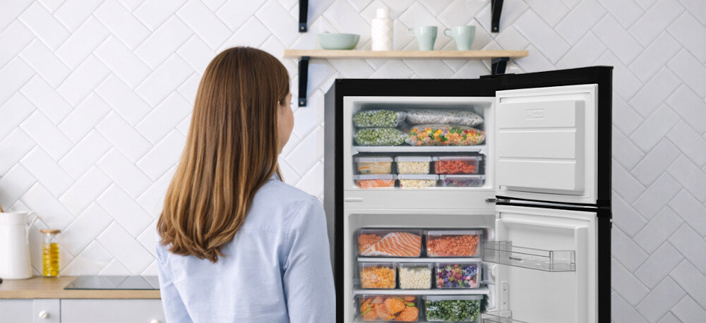 Osoba stoi przed otwartą lodówką wypełnioną różnorodnymi produktami spożywczymi w przezroczystych pojemnikach. W tle widoczna jest biała ściana z półką, na której znajdują się kubki i pojemniki.