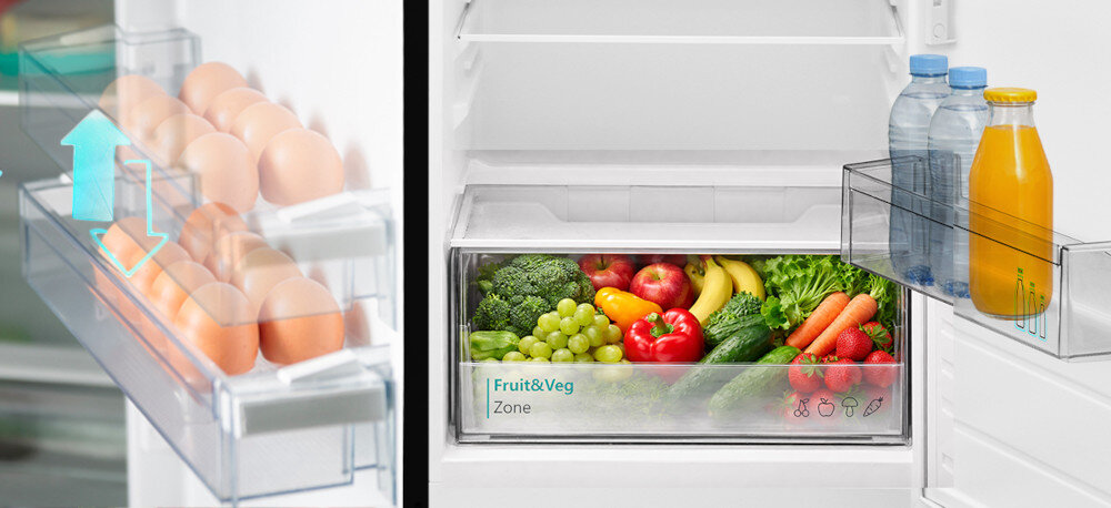 Wnętrze lodówki z widocznymi półkami na jajka oraz szufladą oznaczoną jako \'Fruit & Veg Zone\' z różnymi owocami i warzywami. Na drzwiach lodówki znajdują się butelki z napojami.