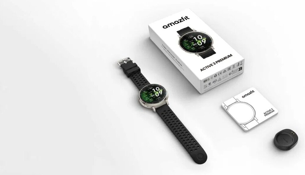 Na obrazie znajduje się smartwatch z czarnym paskiem i okrągłym wyświetlaczem, obok którego leży pudełko z napisem \'amazfit ACTIVE 3 PREMIUM\'. W tle widoczne są instrukcja obsługi i małe akcesorium.