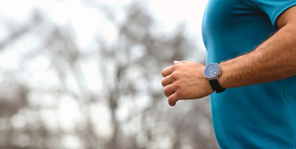 Osoba w niebieskiej koszulce biegnie na zewnątrz, nosząc smartwatch na nadgarstku. W tle widać rozmyte drzewa.