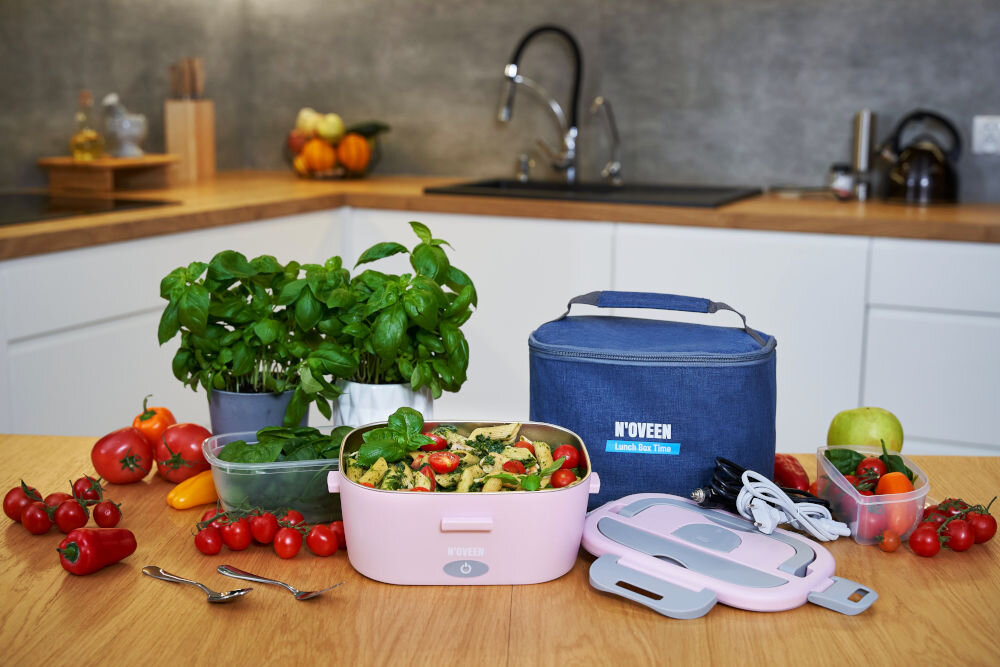 Na blacie kuchennym znajduje się różowy elektryczny lunch box z jedzeniem, obok którego leży niebieska torba z napisem \'N\'OVEEN Lunch Box, Home\'. Wokół rozłożone są świeże warzywa, zioła oraz akcesoria kuchenne.