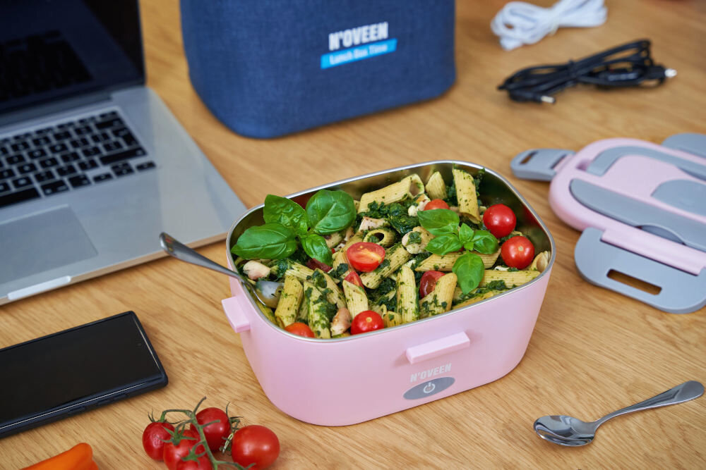 Na drewnianym stole znajduje się różowy elektryczny lunch box NOVEEN z makaronem, pomidorkami koktajlowymi i liśćmi bazylii. W tle widoczny jest laptop, telefon, kabel oraz torba z napisem \'NOVEEN\'.